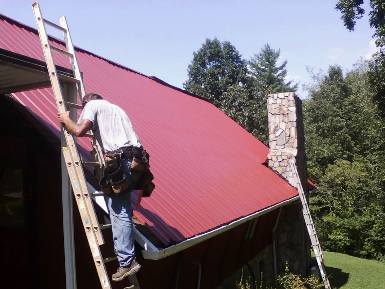 Roofer on a ladder beside a red metal roof and stone chimney — Soffit & Fascia Repair by Roofing Brothers in Armonk